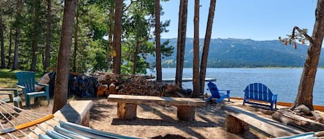 A hammock, a wooden bench, and two blue Adirondack chairs by a lakeside with trees and a distant mountain view.