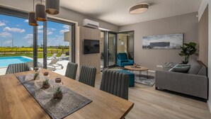 Ein modernes Wohnzimmer mit Esstisch aus Holz, grauen Sofas und einem großen Fenster mit Blick auf einen Swimmingpool.