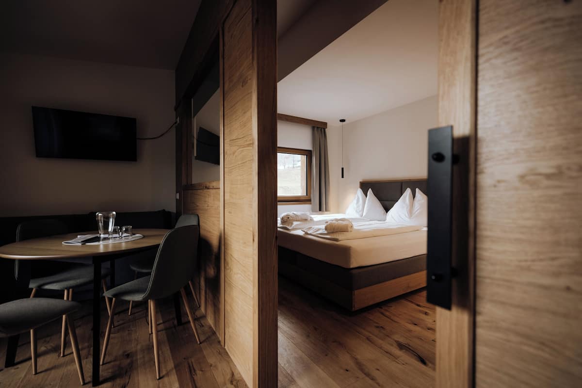 Habitación de hotel con piso de madera, cama con ropa blanca, mesita de noche con vasos y televisor montado en la pared.