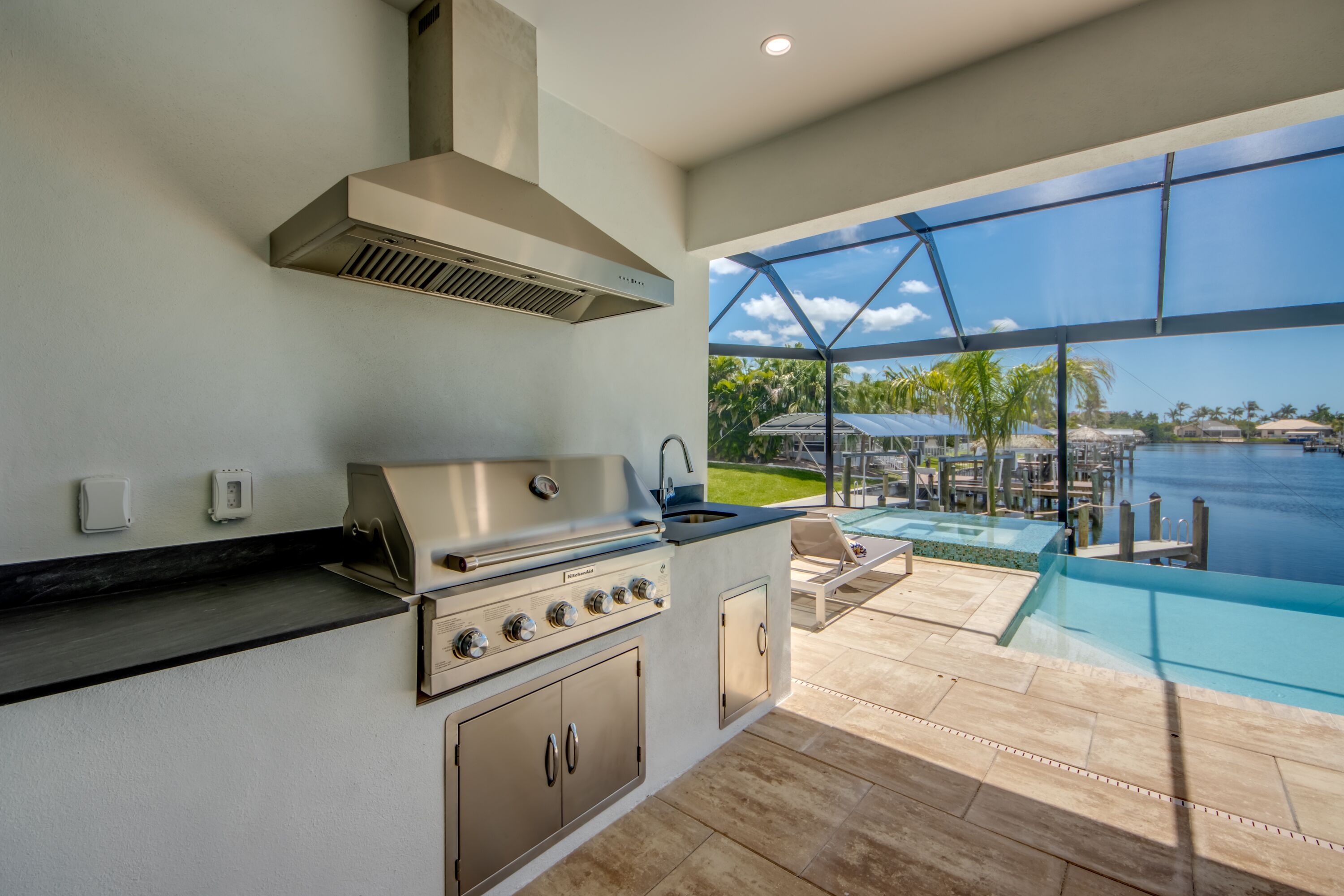 A modern outdoor kitchen with a stainless steel range, a built-in sink, and a large window overlooking a pool and tropical scenery.