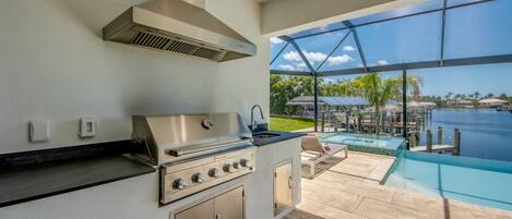 A modern outdoor kitchen with a stainless steel range, a built-in sink, and a large window overlooking a pool and tropical scenery.