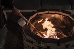 Eine Person bedient ein Feuer in einer runden Feuerstelle im Freien mit einem langen Werkzeug.