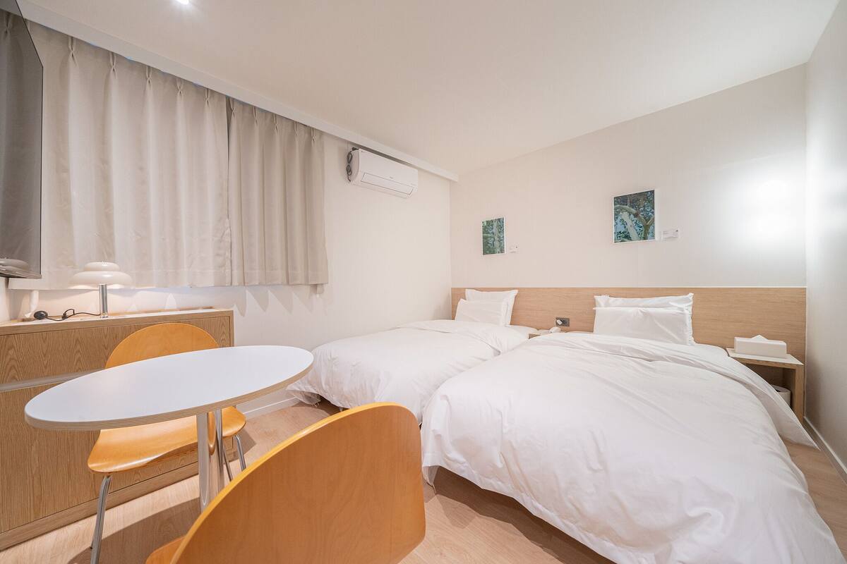 Habitación de hotel con dos camas, un escritorio con silla, una mesa redonda y una ventana con cortinas.