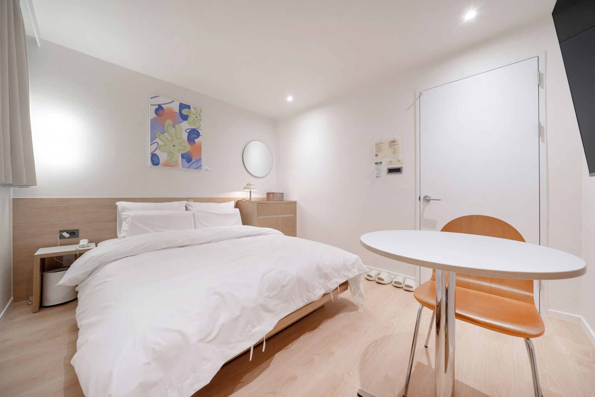 A modern hotel room with a bed, bedside table, lamp, and a round table with a chair.
