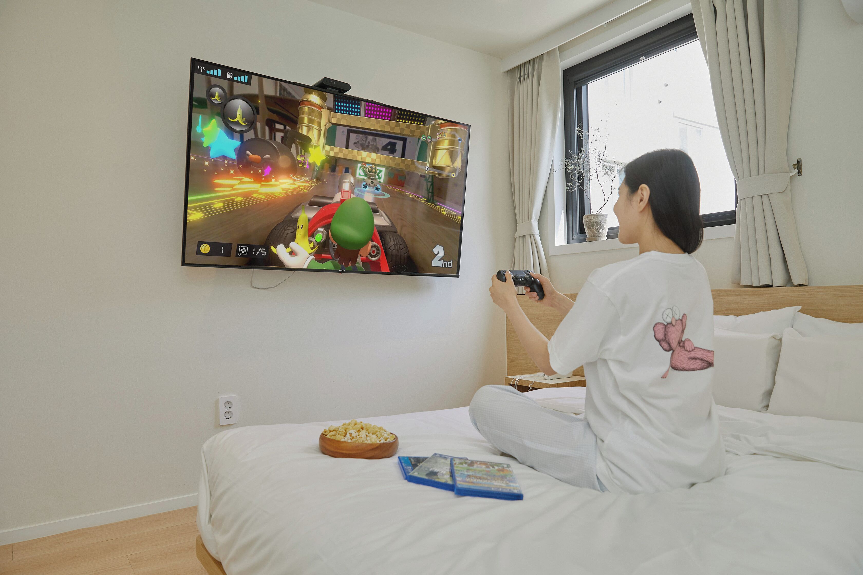Una persona jugando un videojuego en la televisión, en una habitación con cama, un cuenco de palomitas de ma íz y una ventana con cortinas.
