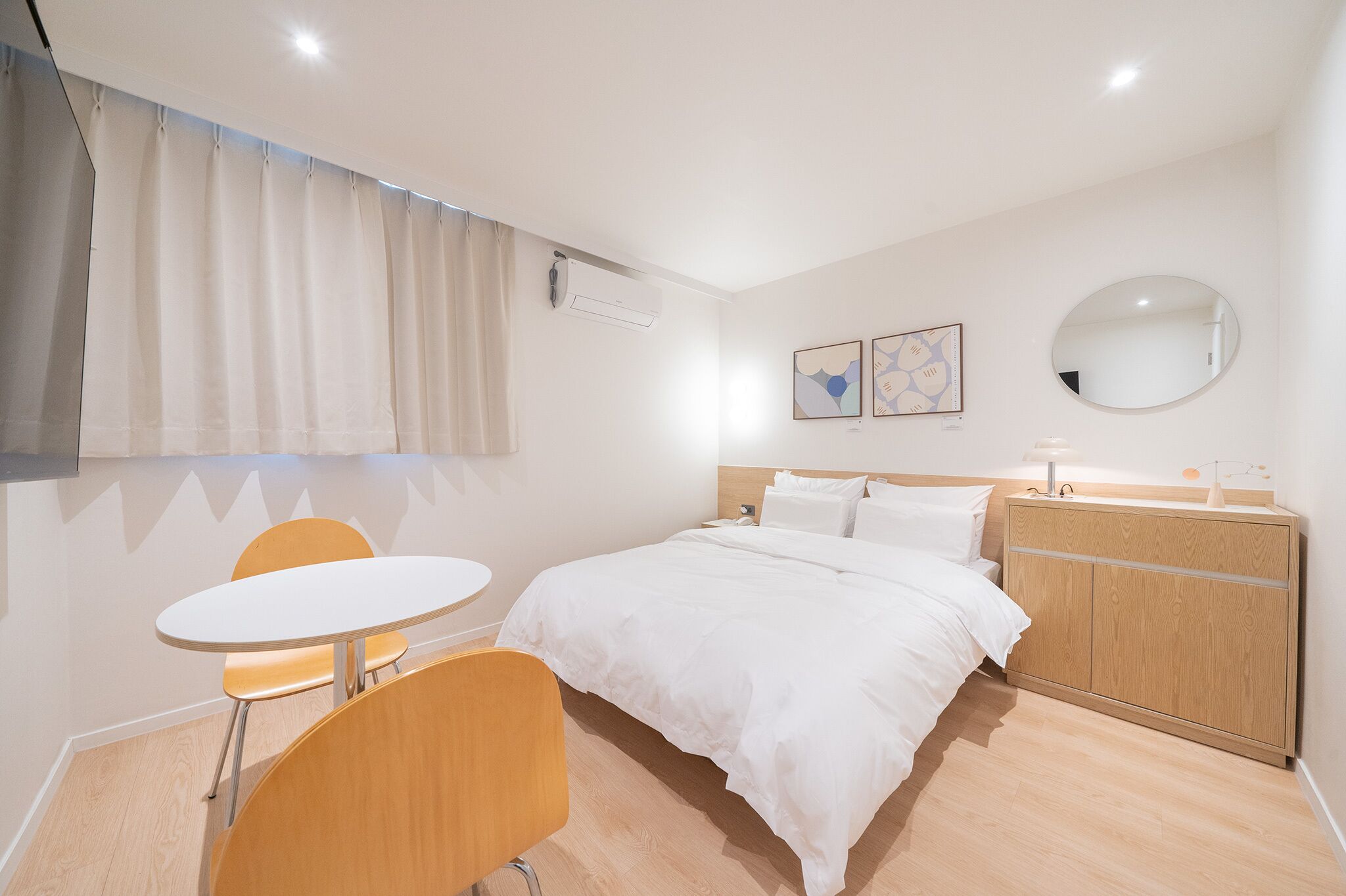Habitación de hotel con cama, espejo redondo, una pequeña mesa con dos sillas y un armario de madera.