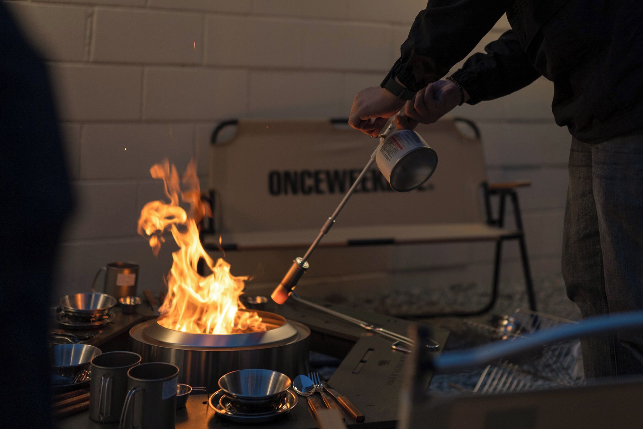 A person cooking over an open flame with a pan, surrounded by cooking utensils and a pot.