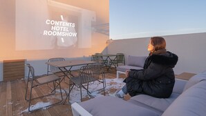Une personne est assise sur un canapé dans une pièce où un écran de projecteur affiche le texte « CONTENU : PRÉSENTATION DES CHAMBRES D’HÔTEL ».