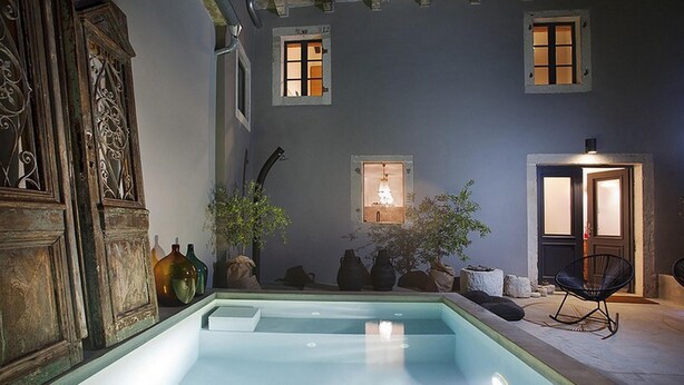Un espace piscine avec une piscine à eau claire, des sièges en pierre et une chaise. On y trouve également des plantes en pot et une porte surmontée d’une lampe.