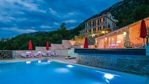 A pool area with lounge chairs and umbrellas, a stone wall, and a building with lights in the background.