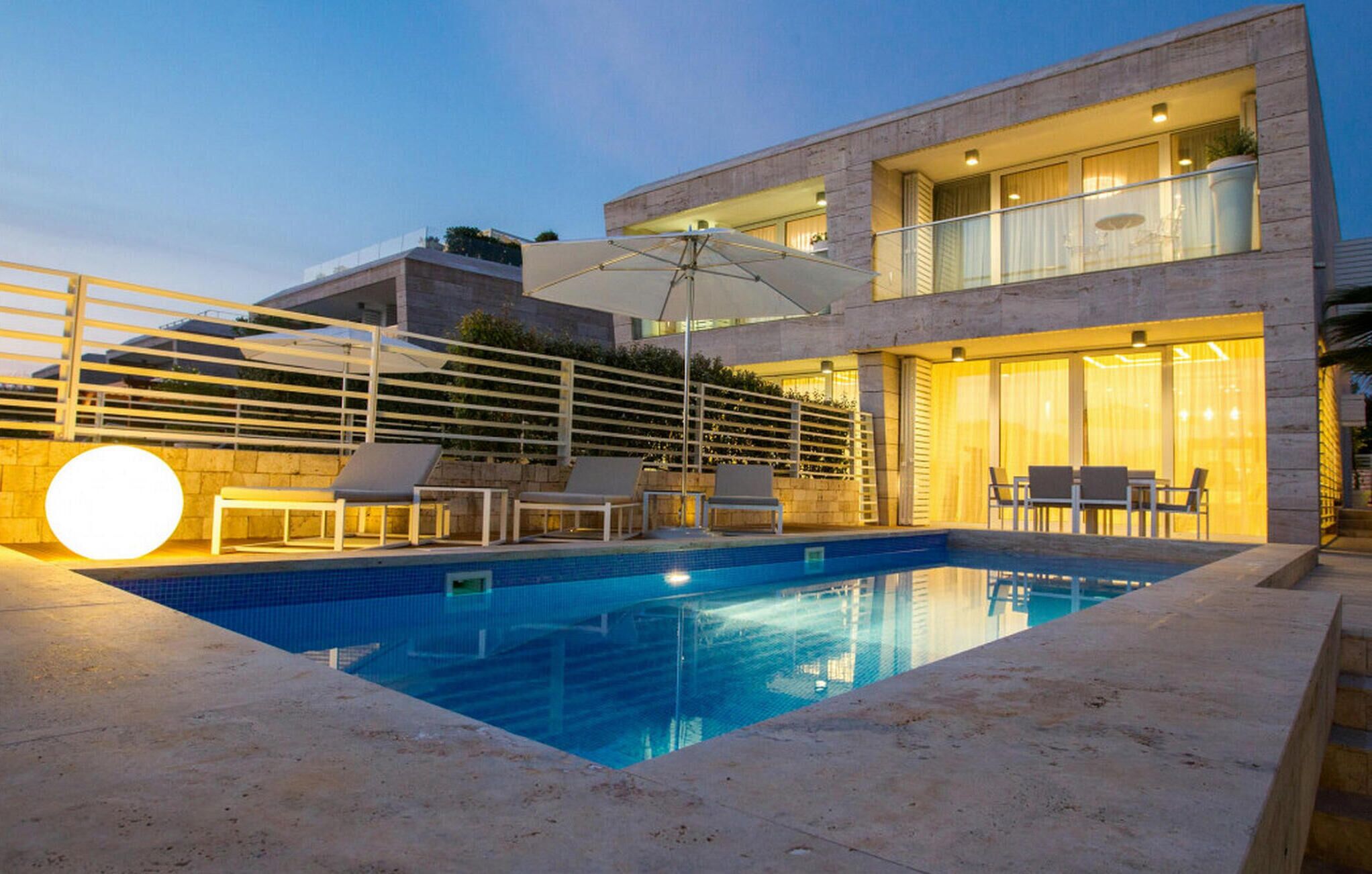 Un área de piscina moderna con piscina cristalina, sillas de descanso y una pérgola con sombrillas.