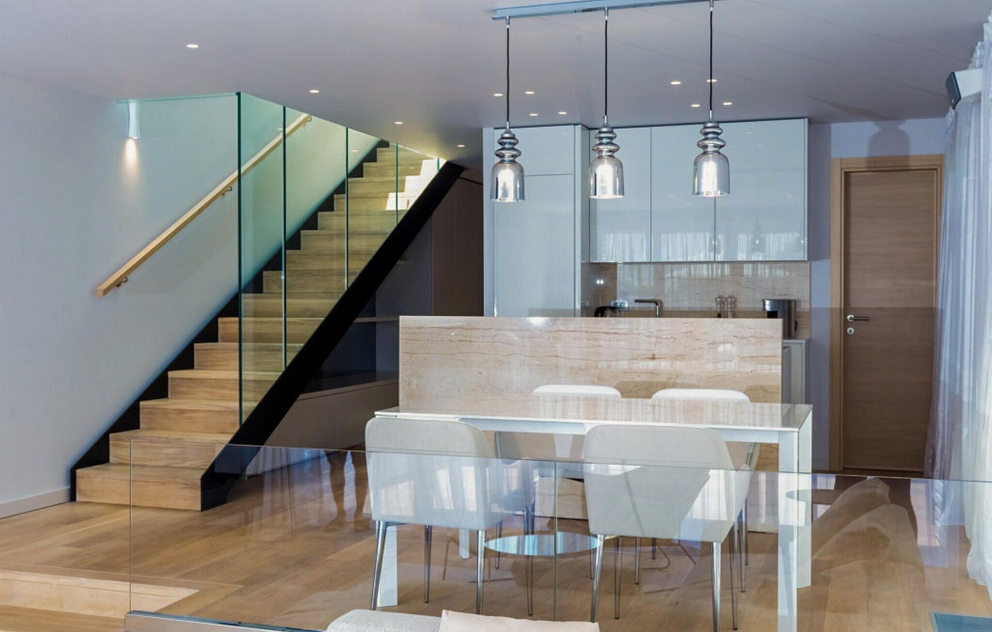 Una cocina moderna con zona de comedor de vidrio, una escalera de madera e iluminación colgante.