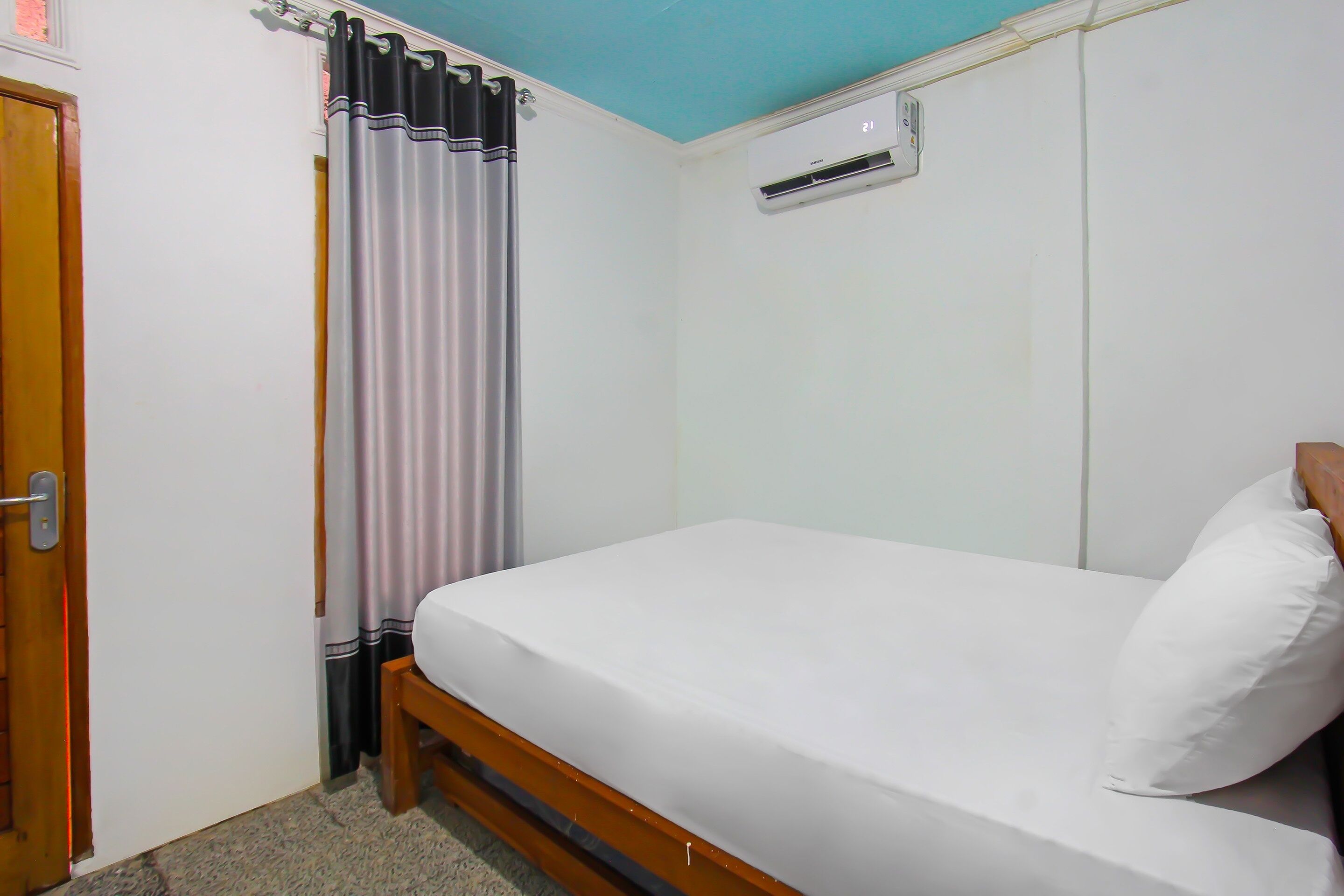 A single bed with a white mattress and pillow, a wooden headboard, a door with a handle, a curtain, and an air conditioning unit on the wall.