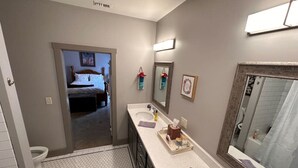Een badkamer met een wit wastafelmeubel, een spiegel en een toilet. Een bed is zichtbaar in de aangrenzende kamer.