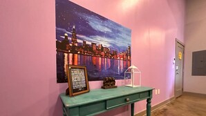 Een smaragdgroene console tafel met een lijst met een bordje en een klein sculptuur, voor een roze muur, met een stadsgezicht schilderij erboven.
