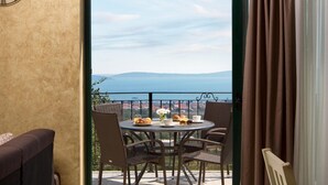 Un balcon avec des fauteuils en osier, une table où sont disposés des aliments pour le petit-déjeuner, et une vue sur la mer et les collines lointaines.