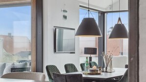 A modern dining area with a large table, four chairs, pendant lighting, and a view of the city through large windows.