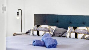 Una cama bien hecha con una toalla azul, dos almohadas estampadas y una lámpara de noche moderna.
