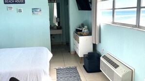A room with a bed, a desk, a chair, a window with blinds, a wall-mounted TV, and a sign reading "BEACH HOUSE". - Salty ~ Tides Studio Condo near the Beach  (Daytona Beach)