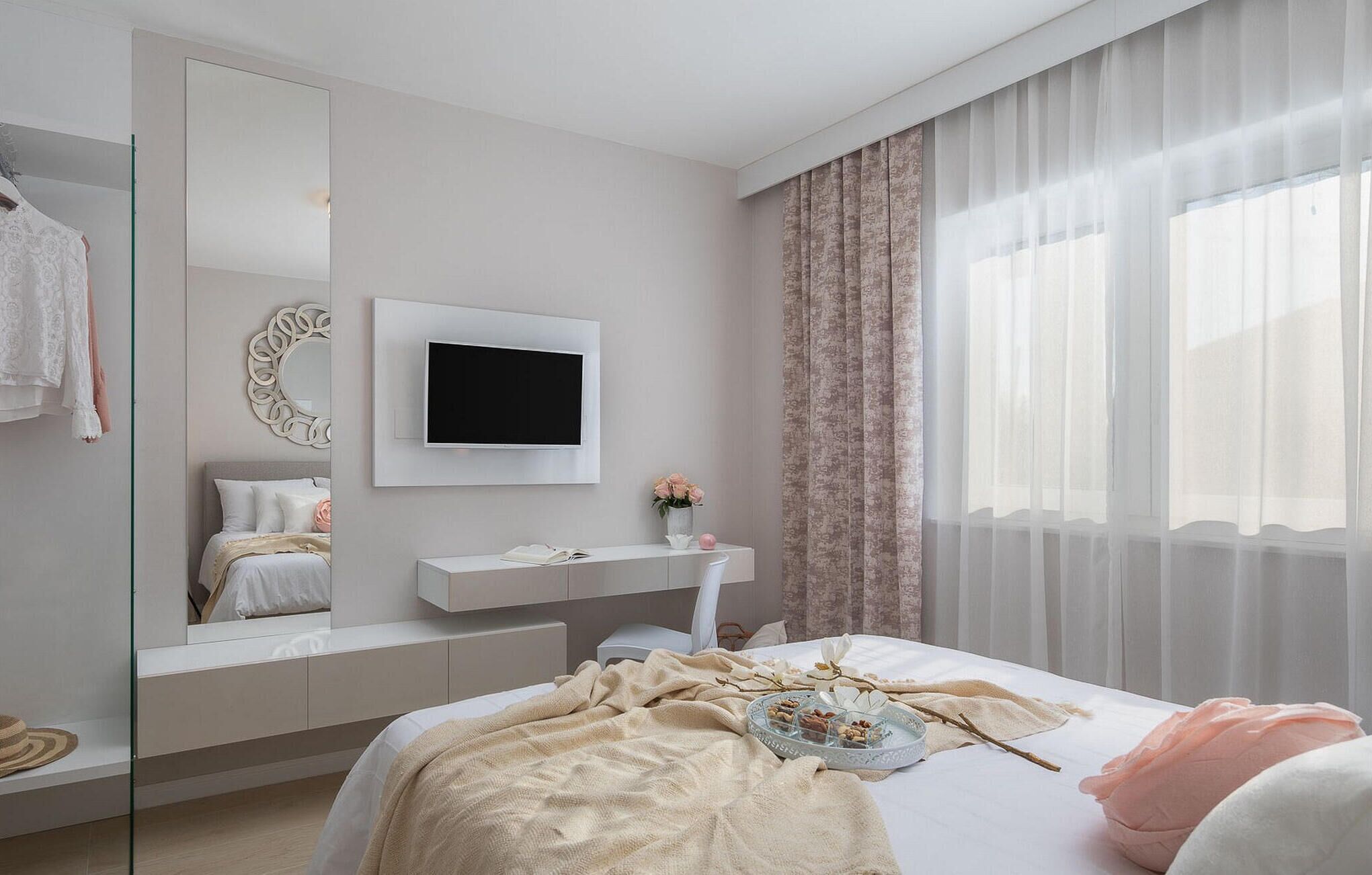 A modern bedroom with a bed, bedside table, mirror, TV, and a window with curtains.