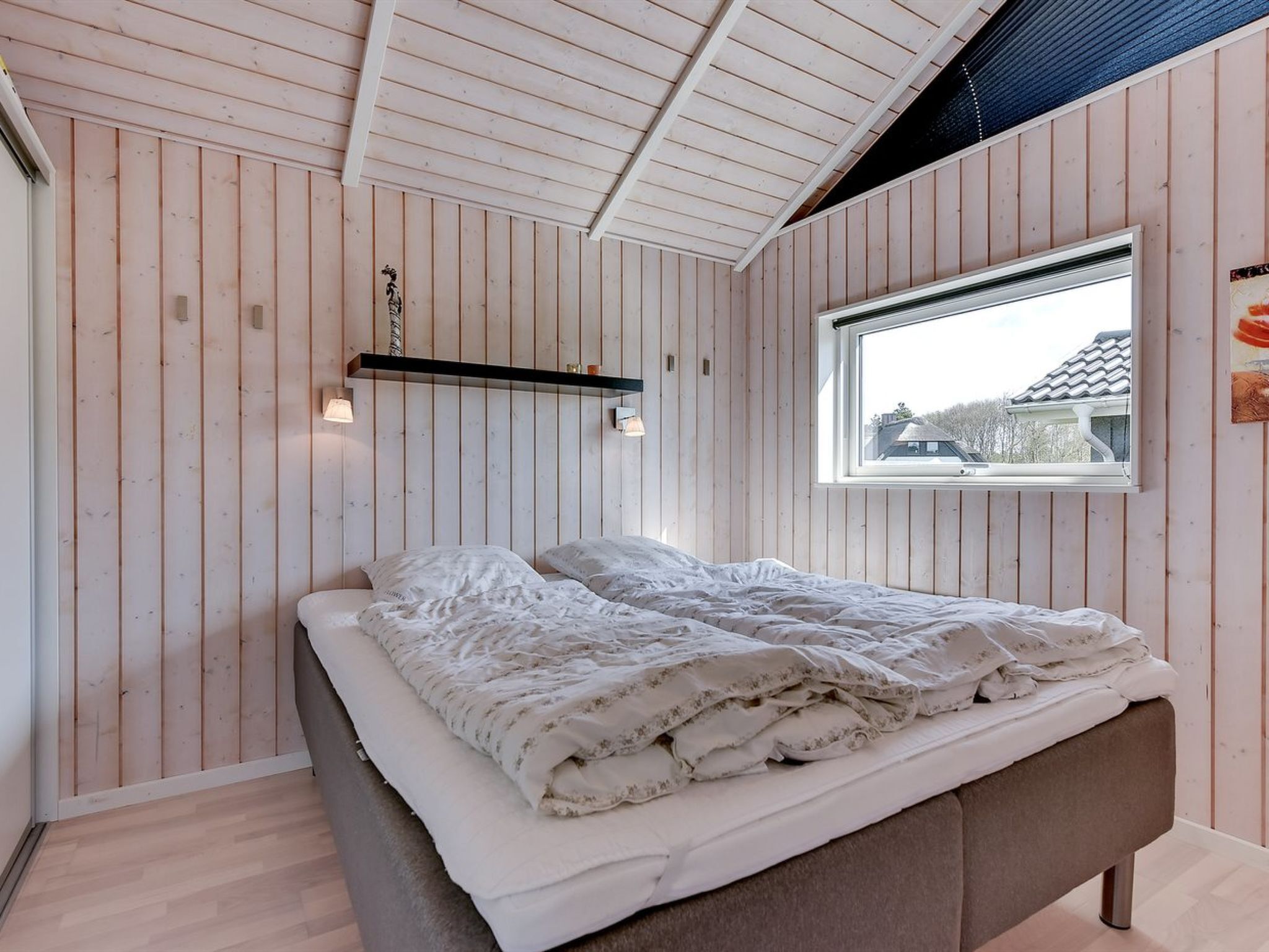 Een slaapkamer met een bed, houten wanden, een raam en een plank met een lamp.