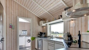 Une cuisine moderne avec des murs en bois, un lave-linge encastré, une grande fenêtre et un plan de travail orné de verres à vin et d’une bouteille.