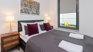 Un dormitorio moderno con una cama grande, dos mesitas de noche con lámparas, una ventana con vistas y un cuadro abstracto de colores en la pared.