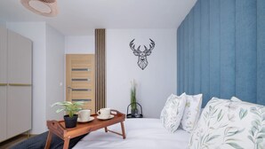 Ein Schlafzimmer mit einem Bett, einem Nachttisch aus Holz mit einer Ablage, einer Topfpflanze und einem an der Wand befestigten Hirsch-Dekoration.
