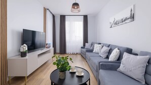 Ein modernes Wohnzimmer mit einem grauen Sofa, einem Fernseher auf einer weißen Konsole, einem schwarzen Couchtisch mit einer Pflanze und einem Stadtbild an der Wand.