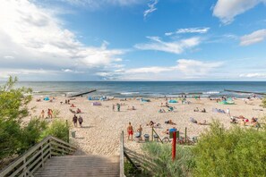 Una playa con una escalera de madera que baja a la arena, un muelle que se adentra en el agua, y gente relajándose en la playa.