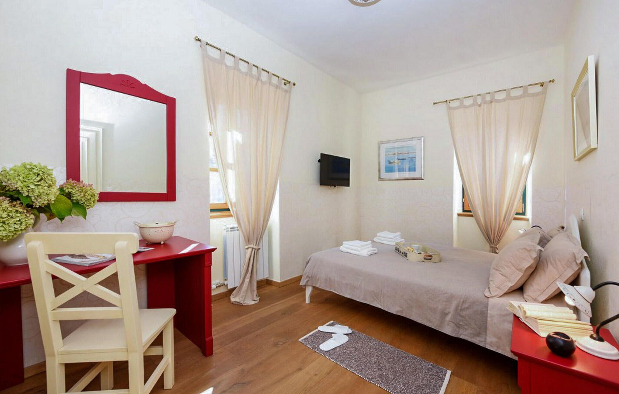 A bedroom with a bed, a desk, a chair, a TV, and a window with curtains.