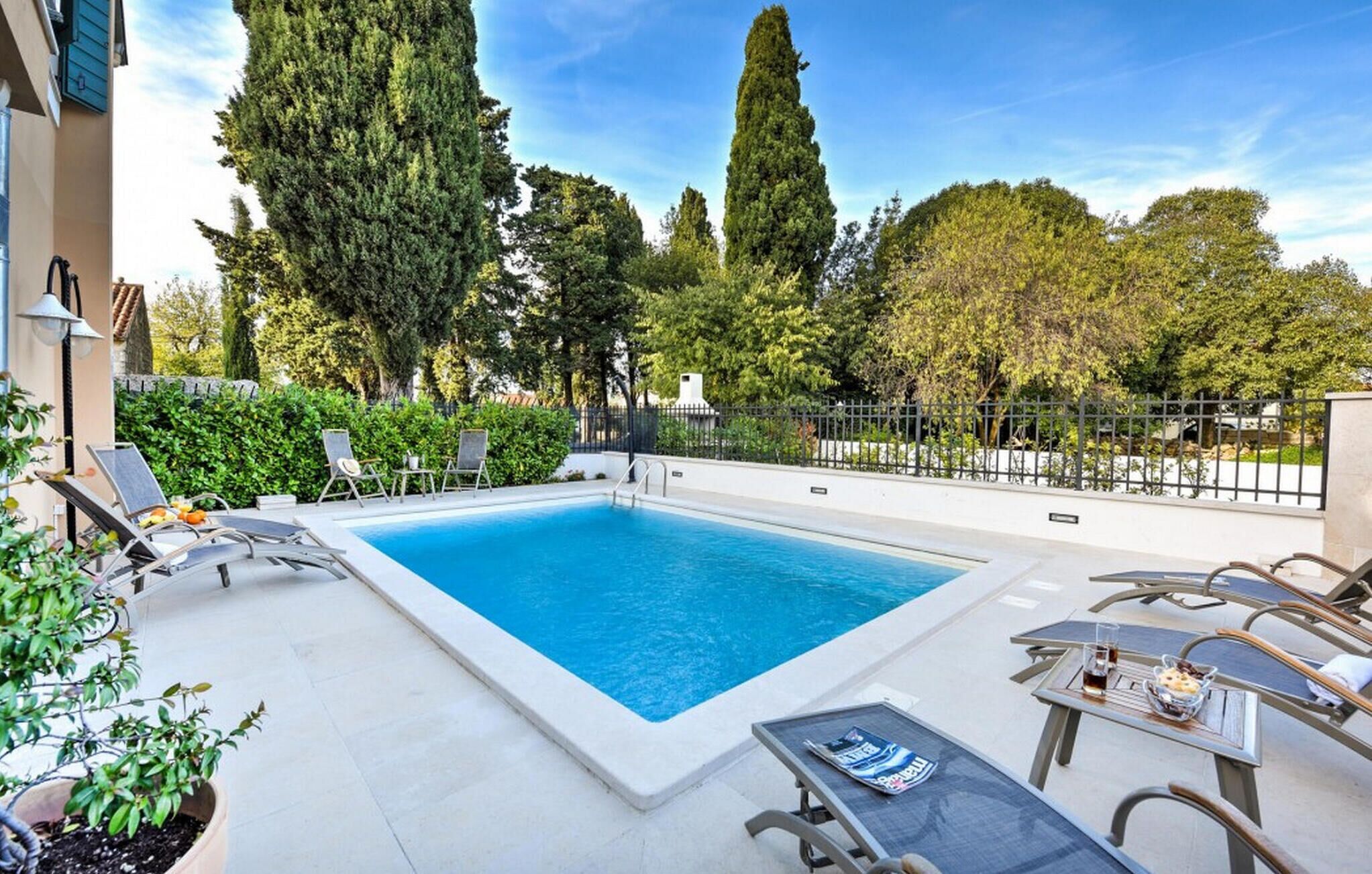 A rectangular swimming pool with lounge chairs and tables around it, surrounded by trees and a fence.