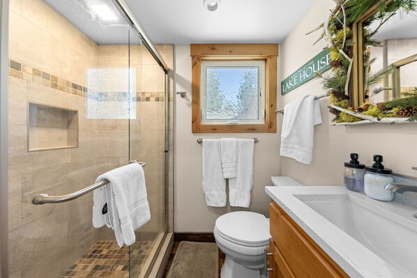 Een badkamer met een douchecabine van glas, een toilet, een wastafel met spiegel, en een raam met uitzicht op bomen.