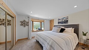 Une chambre à coucher avec un grand lit, une fenêtre avec des rideaux, une tête de lit en bois, un tableau accroché au mur et une petite table de chevet avec une plante.