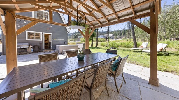 Une terrasse extérieure couverte, avec une table en bois et des chaises en osier, un barbecue et une vue sur une maison et des arbres.
