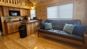 A cabin interior with a wooden floor, a sofa with cushions, a kitchen area with wooden cabinets, a microwave, and a window with blinds. - Surf City Family Getaway | Cabin w/ Loft & Pets OK (Holly Ridge)