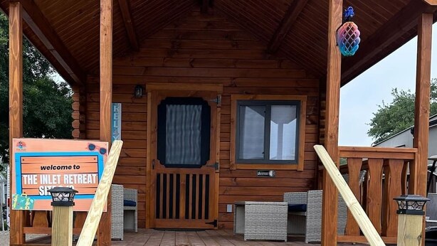 A wooden cabin with a porch, a welcome sign, and a small window. - Surf City Family Getaway | Cabin w/ Loft & Pets OK (Holly Ridge)