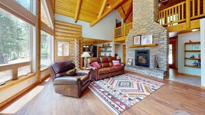 A spacious living room with a stone fireplace, leather furniture, and large windows overlooking a forest. - Log Cabin | WiFi | Secluded 5 Acre Lot | Hoskison Hideaway (Angel Fire)