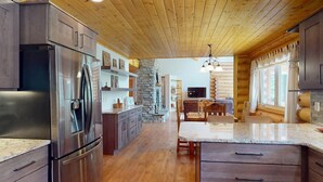 A modern kitchen with a stone fireplace, wooden ceiling, and a central island. - Log Cabin | WiFi | Secluded 5 Acre Lot | Hoskison Hideaway (Angel Fire)