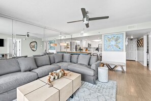 Una sala de estar moderna con un sofá modular, una mesa de centro, un ventilador de techo y una cocina al fondo.