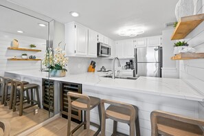 Una cocina moderna con armarios blancos, un refrigerador de acero inoxidable, un microondas y un lavamanos. Hay taburetes de madera y un jarrón con flores en la encimera.