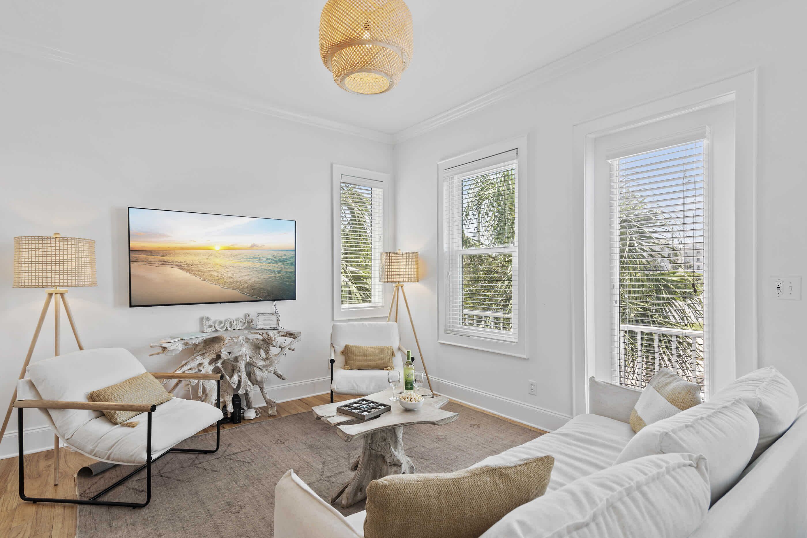 A living room with a white sofa, a white armchair, a coffee table, a TV, and large windows with blinds.