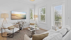 A living room with a white sofa, a white armchair, a coffee table, a TV, and large windows with blinds. - Ocean Views + Walk to Beach + Golf Cart Included! (Santa Rosa Beach)