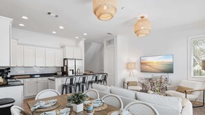 A modern kitchen with white cabinets, a dining area with a wooden table set for four, and a living area with a sofa and chairs. - Ocean Views + Walk to Beach + Golf Cart Included! (Santa Rosa Beach)