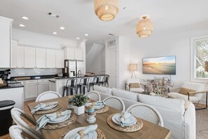 A modern kitchen with white cabinets, a dining area with a wooden table set for four, and a living area with a sofa and chairs.