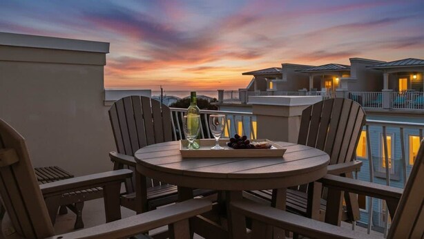 A round table with chairs on a balcony overlooking a swimming pool and adjacent buildings during sunset. - Gulf Views | Rooftop Patio | Heated Pool (Santa Rosa Beach)