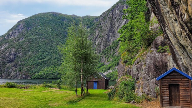 A cabin with a blue roof and a wooden door, surrounded by a grassy area and a rocky cliff.