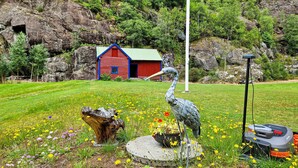 A red building with a green roof, a large bird statue, a tree stump, and a lawn with yellow flowers.