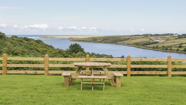 Una mesa de picnic con bancos en un área con césped, con vistas a un cuerpo de agua y colinas distantes.