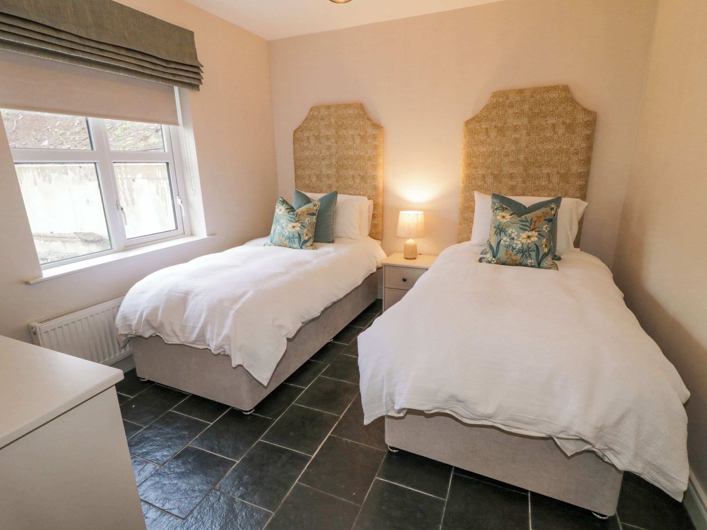 A twin bedroom with two beds, a window, a radiator, and a nightstand with a lamp.